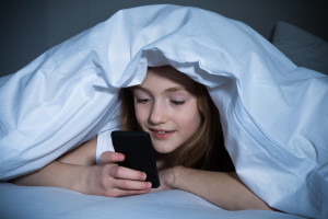 Happy Girl Looking At Mobile Phone While Lying Under Blanket On The Bed